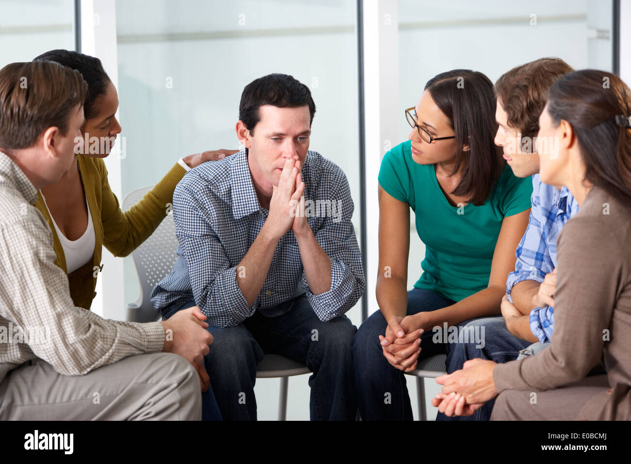 A support group closely circled around a man with his hands pressed to his nose and mouth, palms touching.  He looks sad and the 5 people around him look supportive.