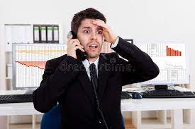 A man in a suit in an office who is on the phone, looking shocked and worried. His left hand is slapping his head.